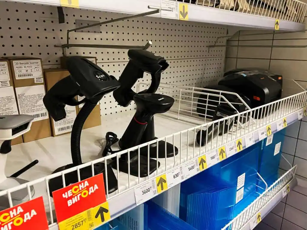 Barcode scanners and related electronic devices are displayed on a store shelf with price tags. Some items are in boxes, while others are out for viewing. Yellow sale signs are visible below the items.