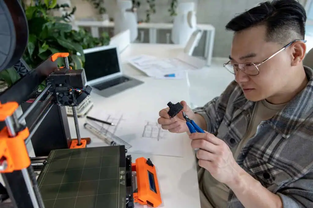 A person wearing glasses examines a 3D-printed part with pliers at a desk, next to a 3D printer, a laptop, and technical drawings in a modern, bright workspace.