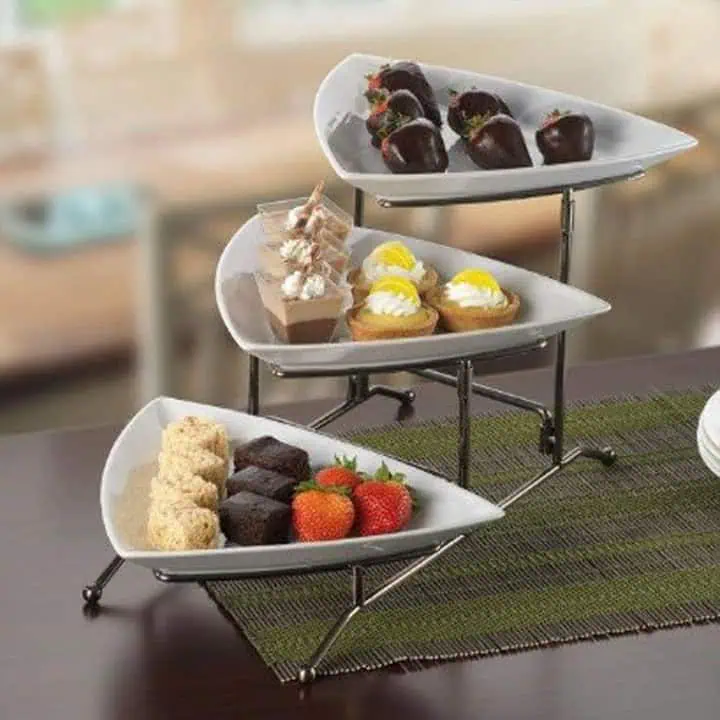Three-tiered serving stand with white, leaf-shaped plates displaying assorted desserts: chocolate-covered strawberries, mini parfaits, lemon tarts, cake squares, brownies, and fresh strawberries.