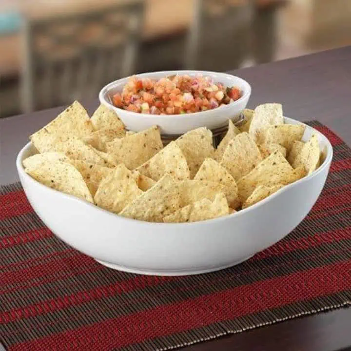 A large white bowl filled with tortilla chips, with a smaller bowl of chunky salsa resting inside it, placed on a red and brown woven placemat on a table.