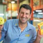 A smiling man in a blue Scrub Daddy shirt holds a yellow smiley-face sponge in one hand and a Scrub Daddy box in the other, standing in a warehouse setting.