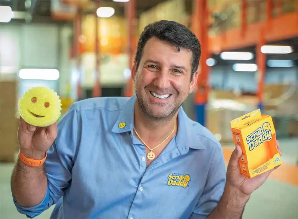 A smiling man in a blue Scrub Daddy shirt holds a yellow smiley-face sponge in one hand and a Scrub Daddy box in the other, standing in a warehouse setting.
