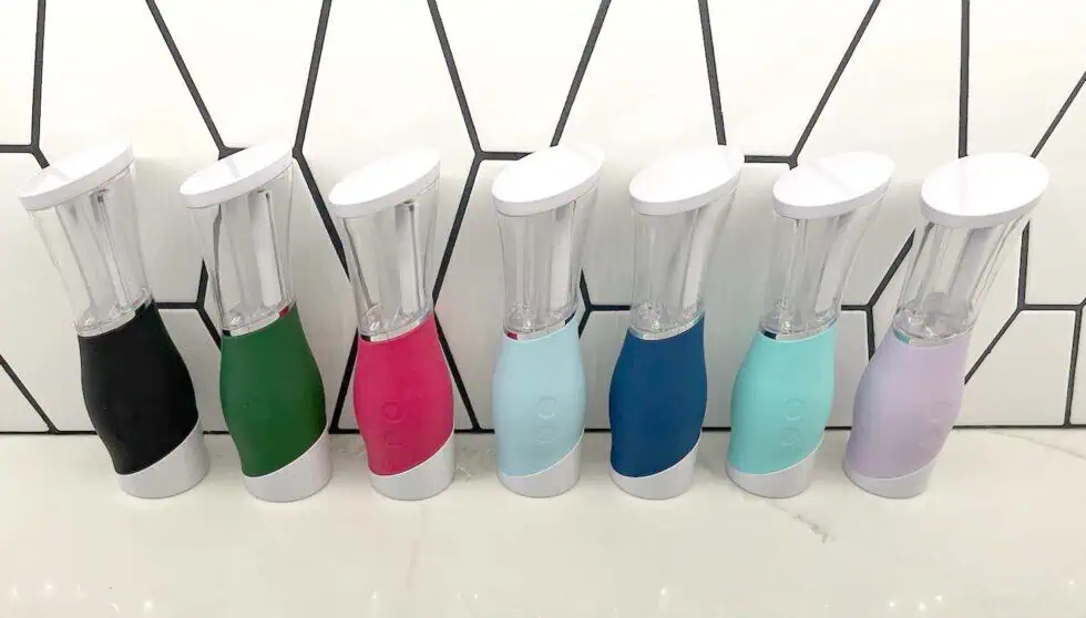 Seven colorful electric toothbrushes with white tops are lined up on a white countertop in front of a white tile wall with black geometric lines. The toothbrush bases are in black, green, pink, light blue, turquoise, blue, and lavender.