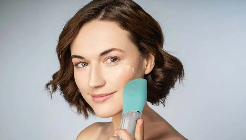 A woman with short brown hair smiles while holding a blue facial cleansing device to her cheek, with a small amount of cream visible on her skin, against a light background.