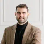 A man with short brown hair and a beard wears a tan blazer over a black turtleneck, smiling slightly while standing in front of a white paneled wall.