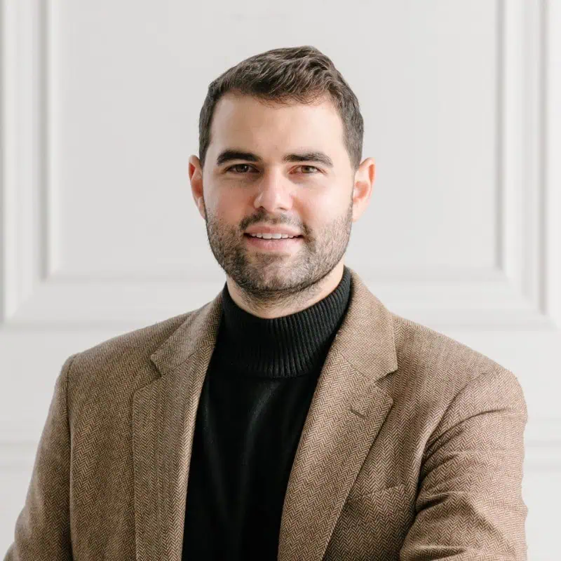 A man with short brown hair and a beard wears a tan blazer over a black turtleneck, smiling slightly while standing in front of a white paneled wall.