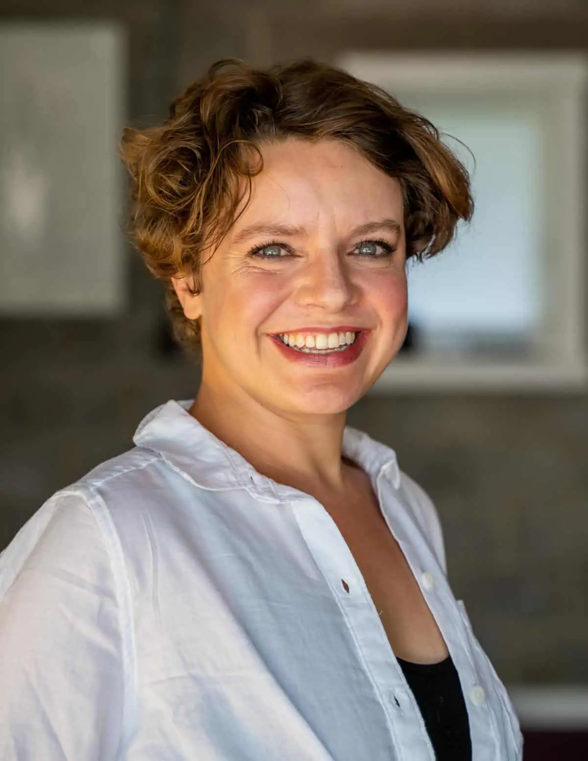 A woman with short, curly brown hair smiles warmly at the camera. She is wearing a white button-up shirt over a black top, standing indoors with a blurred background.