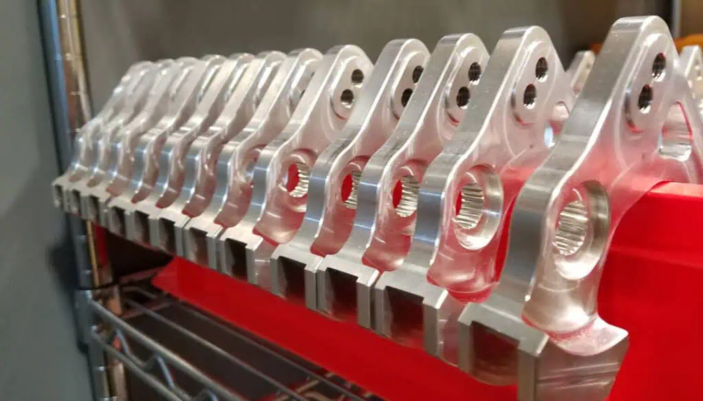 A row of identical silver metal parts with holes and angled edges are lined up on a red tray on a metal shelf, likely in a workshop or manufacturing setting.