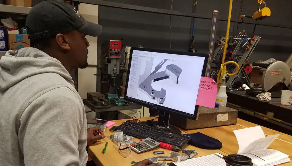 A person wearing a hoodie and cap sits at a desk, looking at a computer screen displaying a 3D CAD model in a workshop. Tools, notes, and papers are scattered on the wooden workbench.