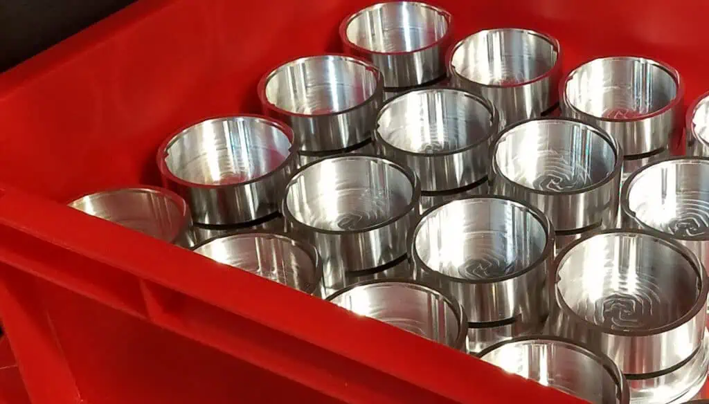 A red plastic bin filled with shiny, cylindrical metal parts arranged in neat rows, reflecting light from their smooth, polished surfaces.