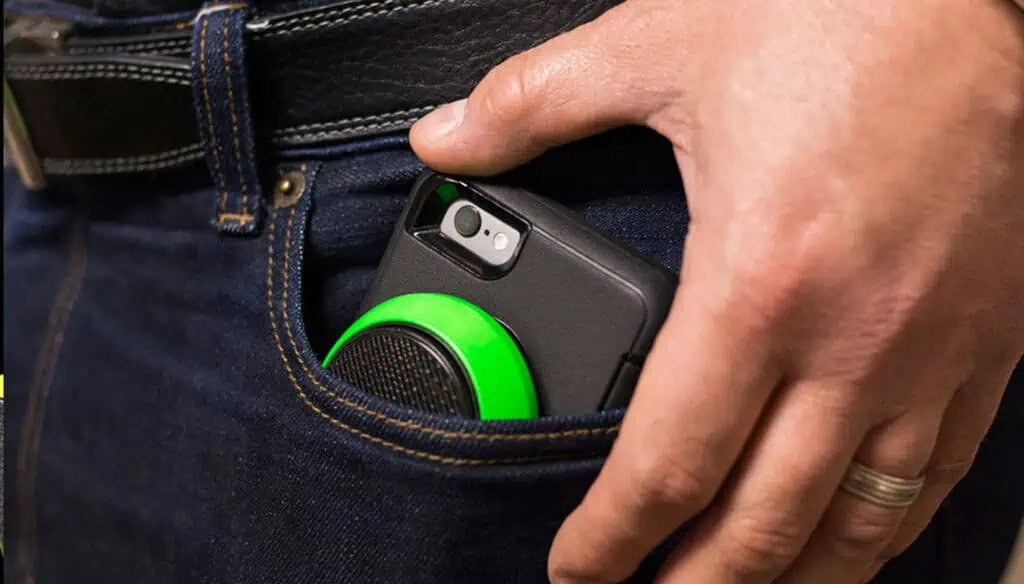 A person is putting a smartphone with a black case and a round green attachment into the front pocket of dark blue jeans. The person is wearing a black belt and a silver ring on their finger.