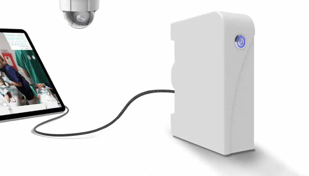 A white electronic device with a glowing power button is connected by a cable to a tablet displaying a video call with a doctor. A security camera is visible above, all set against a white background.