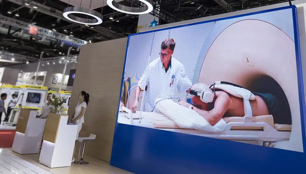 A large digital screen at a medical exhibition displays an image of a doctor assisting a patient in an MRI machine, while a woman stands at a reception booth nearby.