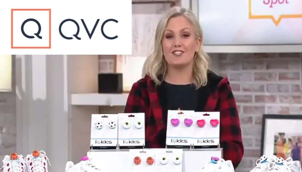 A smiling woman stands behind a display of shoelace charms on a TV shopping set. The QVC logo appears in the top left corner. The background includes a brick wall and shelves with decor.