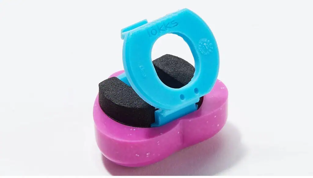 A small, colorful plastic device with a pink base and a blue hinged top, featuring two black foam pads inside, designed for cleaning rings or small jewelry items.