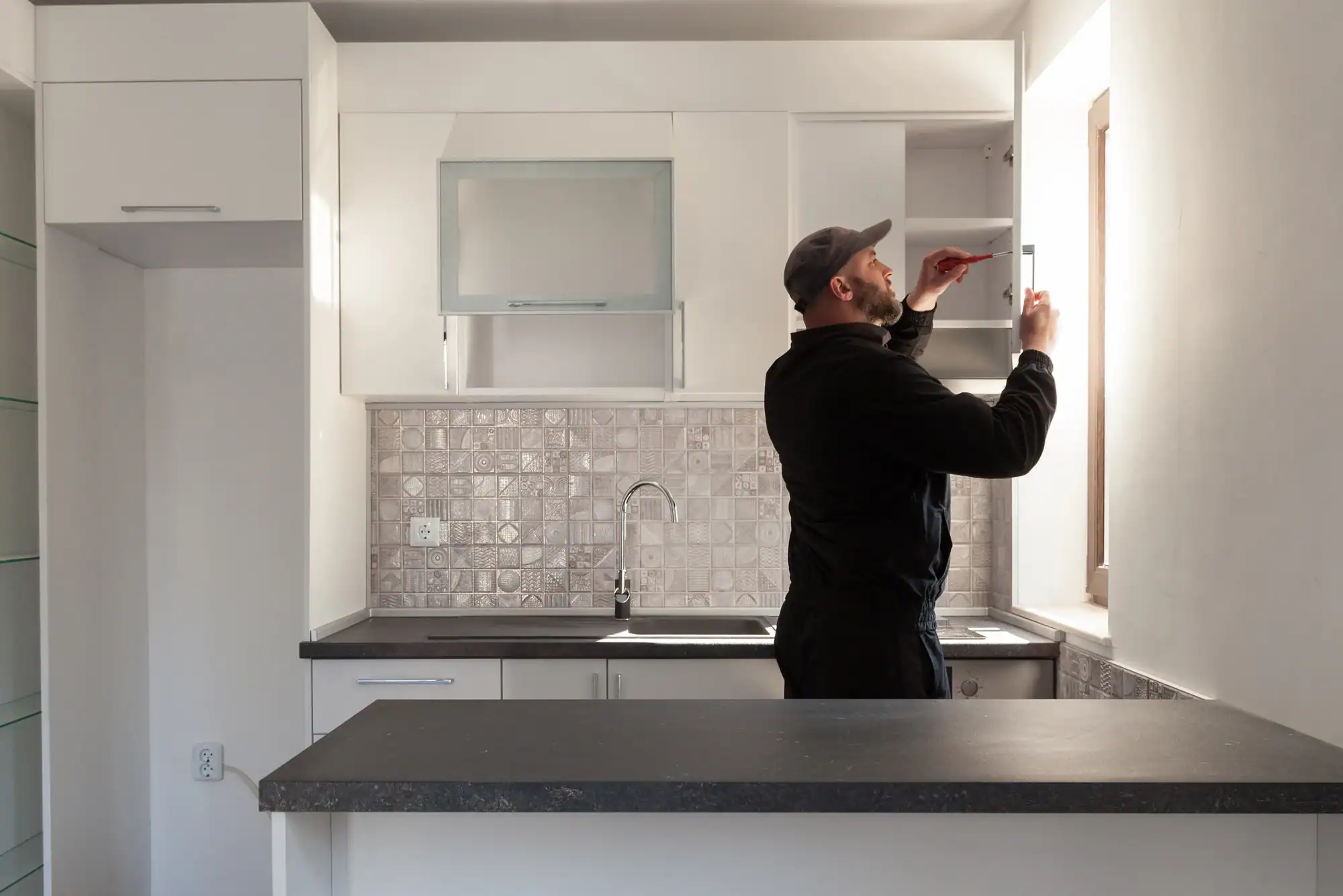 A man in a black outfit and cap uses a screwdriver to fix or install a cabinet in a modern, white kitchen with patterned tile backsplash and dark countertops. Sunlight streams through a window to his right.