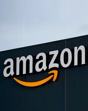 The image shows the Amazon logo on the side of a building, with amazon in large white letters and a yellow arrow forming a smile underneath. The background is a dark exterior wall under a cloudy sky.
