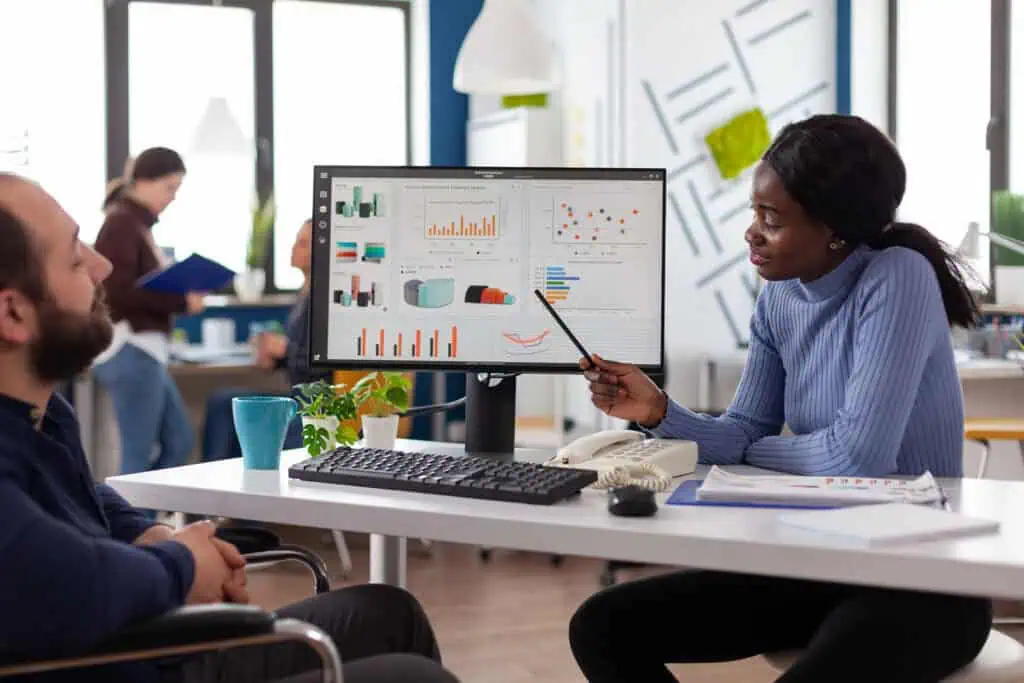 A woman points at a computer screen displaying various charts and graphs, while discussing data with a man seated across from her in a modern office setting.