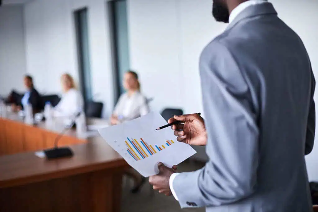 A person in a suit holds a paper with colorful bar graphs and points at it with a pen, while three people sit at a conference table in the background, slightly out of focus.