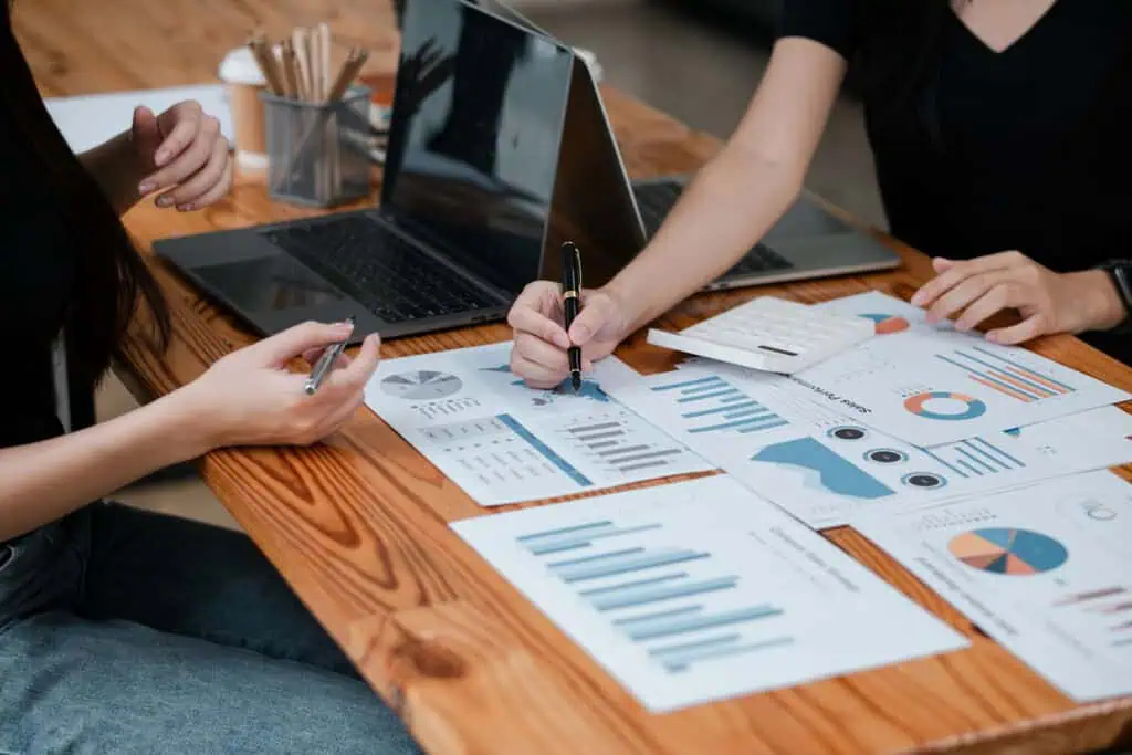 Two people sit at a wooden table reviewing charts and graphs on paper, with a laptop nearby. One person points with a pen at a document, discussing data and analysis.