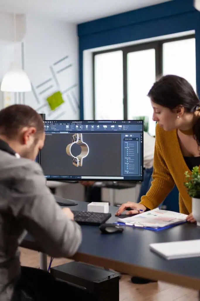A man sits at a desk using a computer to design a watch in 3D software while a woman in a yellow sweater stands beside him, looking at the screen and discussing the project in a modern office.
