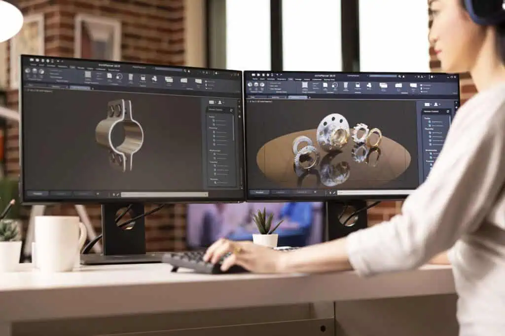 A person works at a desk with two monitors displaying 3D mechanical part designs in CAD software. The workspace includes a keyboard, mouse, coffee mug, and small potted plants.