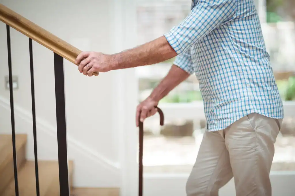 An older adult wearing a blue checkered shirt and beige pants is walking up stairs, holding onto the handrail with one hand and using a wooden cane with the other for support.