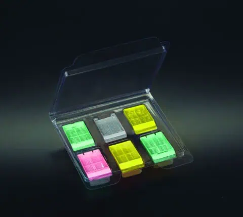 A clear plastic case containing six rectangular biopsy cassettes in green, pink, yellow, and white, arranged in two rows on a black background.