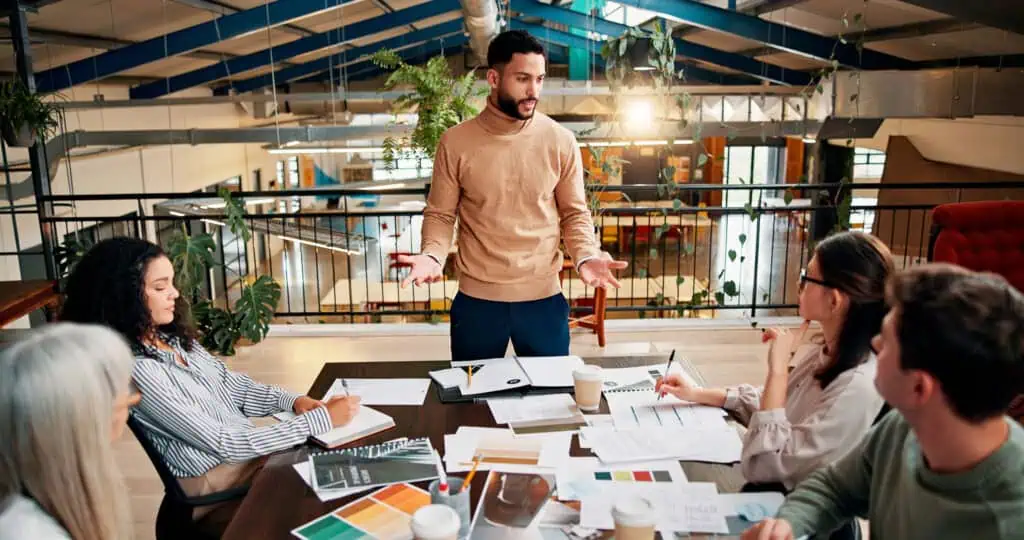 A man stands and speaks to four colleagues seated around a table covered with documents, notebooks, and coffee cups in a modern, open office space with plants and natural light.