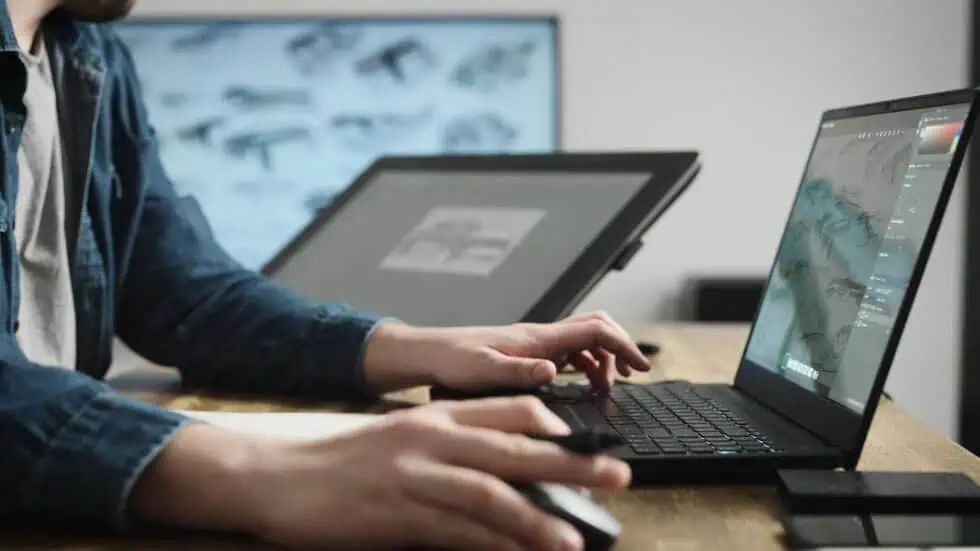 A person using a laptop and a graphics tablet at a desk, working on digital illustrations of firearms. The screen shows sketching software and gun designs in progress.