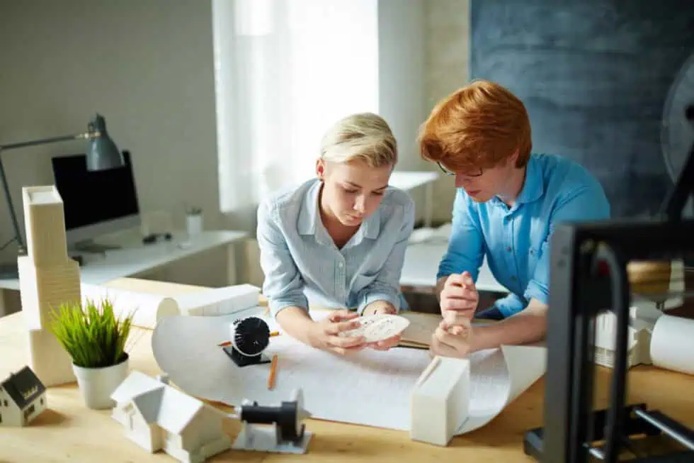 Two people examine a scale model and building plans at a desk in a well-lit office, surrounded by architectural models and design tools.