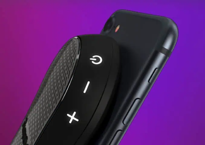 A close-up of a black portable speaker with power and volume buttons resting against the back of a black smartphone, set against a purple gradient background.