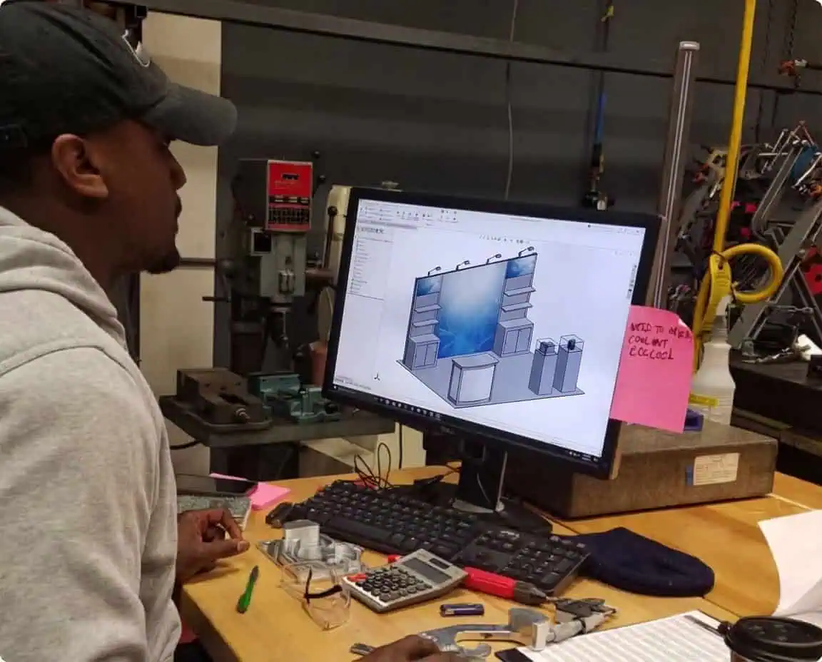 A person wearing a cap and hoodie sits at a desk in a workshop, looking at a desktop monitor displaying a 3D CAD model of an exhibition booth. Various tools and notes are scattered on the desk.