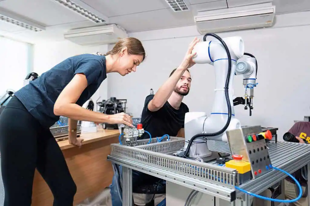 Two people work together in a lab, adjusting a white robotic arm on a metal workbench with control panels and wires, focusing on robotics or automation technology.