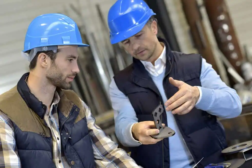Two men wearing blue safety helmets and vests examine a metal part together in an industrial workshop setting. One man explains or points out details while the other listens attentively.