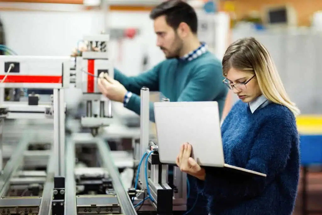 A woman holding a laptop stands in the foreground, focused on her screen, while a man in the background adjusts machinery in a modern industrial or engineering lab setting.