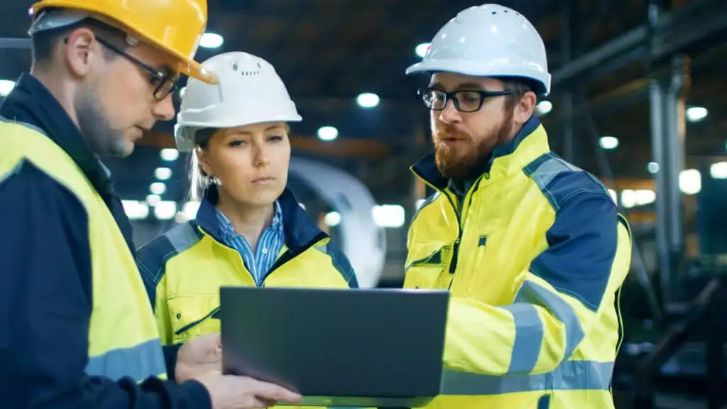 Three construction workers wearing yellow safety jackets and white helmets stand indoors, looking at a laptop. One person gestures towards the screen while discussing something with the others in an industrial setting.