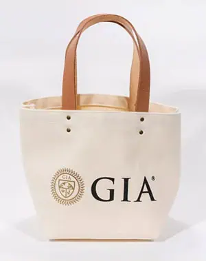 A cream-colored canvas tote bag with brown handles, featuring the GIA logo and emblem printed in black and gold on the front.