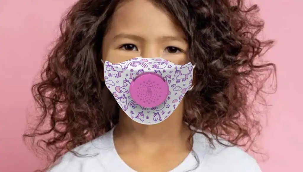 Young child with curly hair wearing a white shirt and a white face mask decorated with purple unicorns and rainbows, standing in front of a pink background.