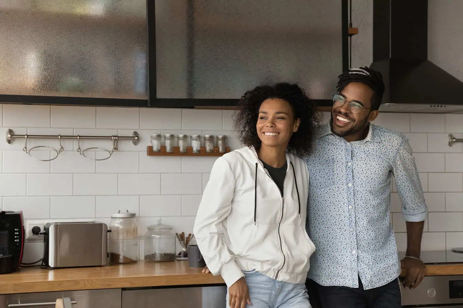 A smiling couple stands together in a modern kitchen, looking off to the side. The woman wears a white hoodie and the man wears glasses and a patterned shirt. Kitchen shelves, jars, and appliances are visible in the background.