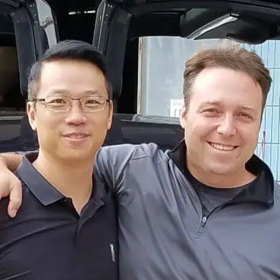 Two men stand close together, smiling at the camera. The man on the left wears glasses and a black polo shirt, while the man on the right wears a gray zip-up jacket. One has his arm around the other.