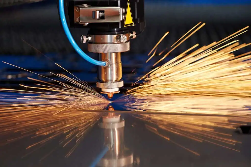 A close-up of a CNC laser cutting machine slicing through a sheet of metal, with bright yellow-orange sparks flying outward from the point of contact.