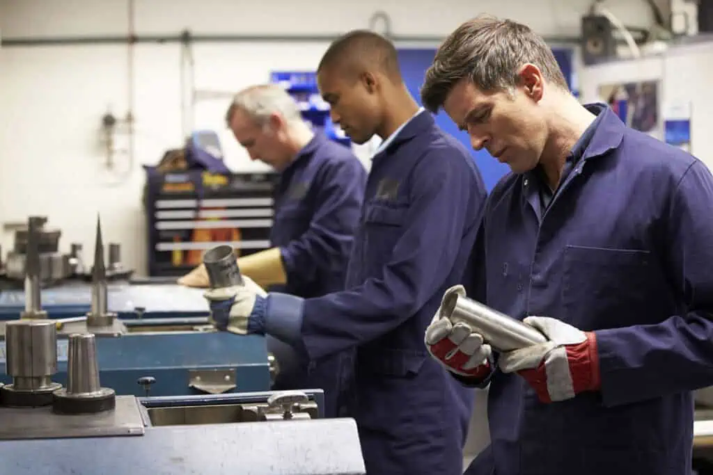 Three men in blue work uniforms operate machinery and inspect metal parts in an industrial workshop. The man in the foreground wears gloves and examines a cylindrical piece.