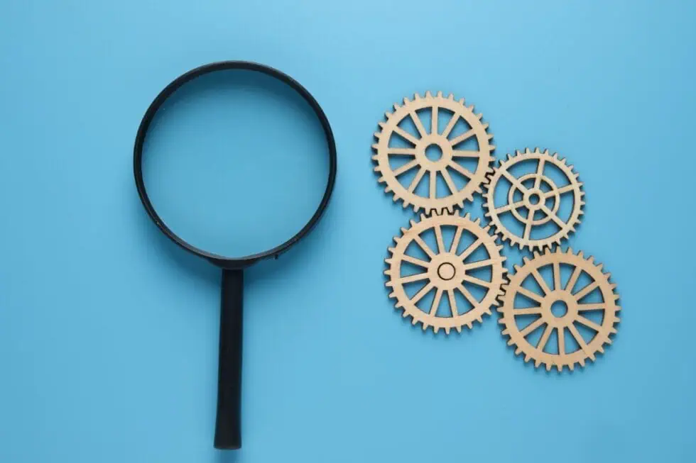 A black magnifying glass lies next to five interlocking wooden gears on a blue background.