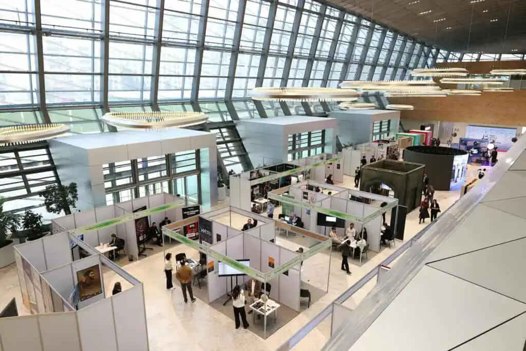 An indoor exhibition hall with modern architecture, tall glass windows, and several booths arranged in rows. People are walking, browsing displays, and interacting at the event.