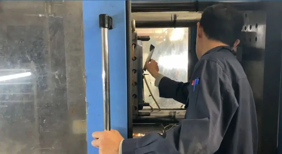 A person in dark work clothes operates machinery, inspecting or adjusting equipment inside an industrial machine with a reflective door and blue frame.
