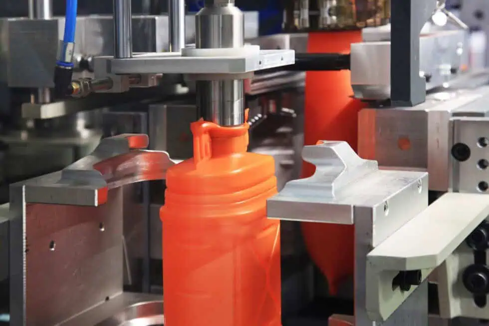 A close-up of an orange plastic bottle being formed by an industrial blow molding machine, with metal clamps and mechanical components visible around the bottle.