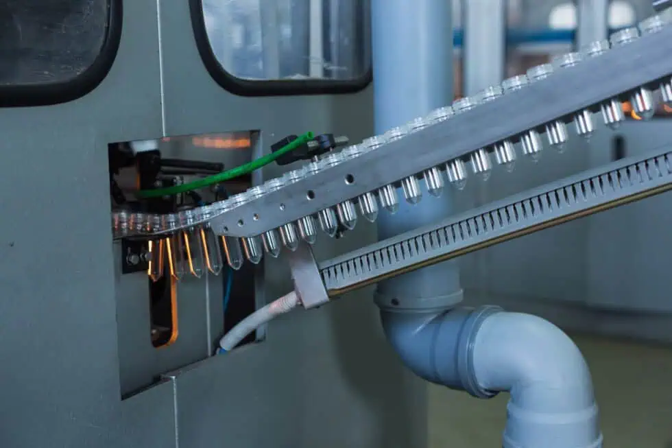 A close-up view of a conveyor system in a factory, transporting transparent plastic bottles through a machine for processing or filling, with pipes and machinery visible in the background.