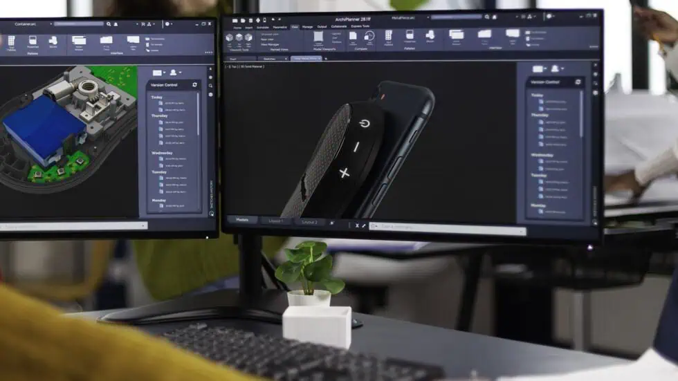 Two computer monitors display 3D design software, one showing a building model with colored sections, the other a detailed image of a smartphone. A small potted plant sits on the desk below the screens.