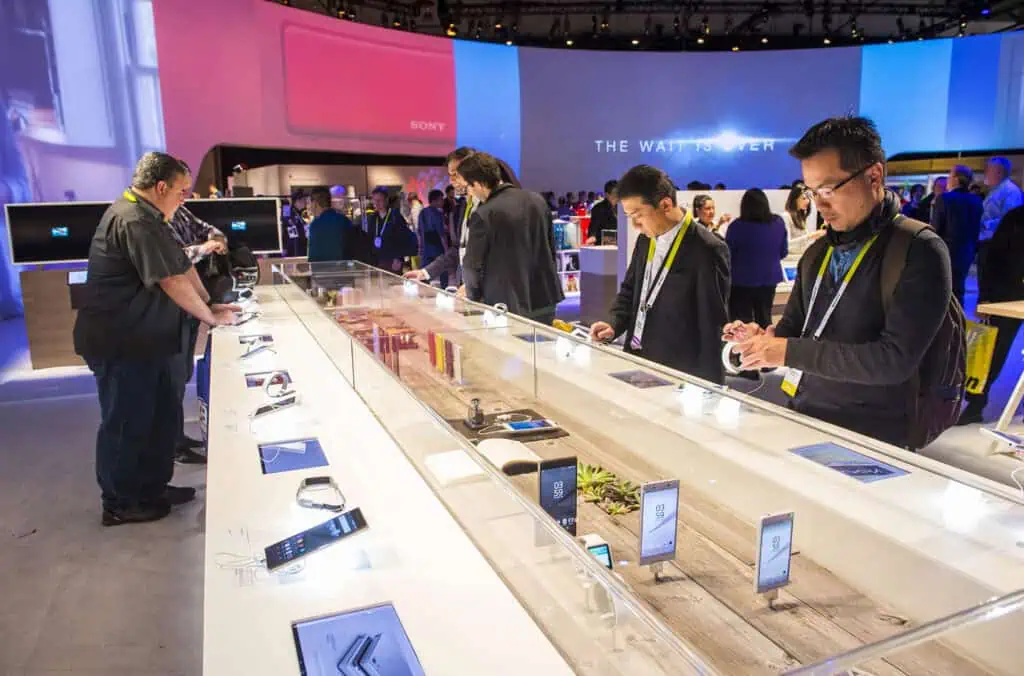 People at a tech expo interact with smartphones and other gadgets displayed on a long counter, surrounded by screens and product displays in a brightly lit, modern exhibition hall.